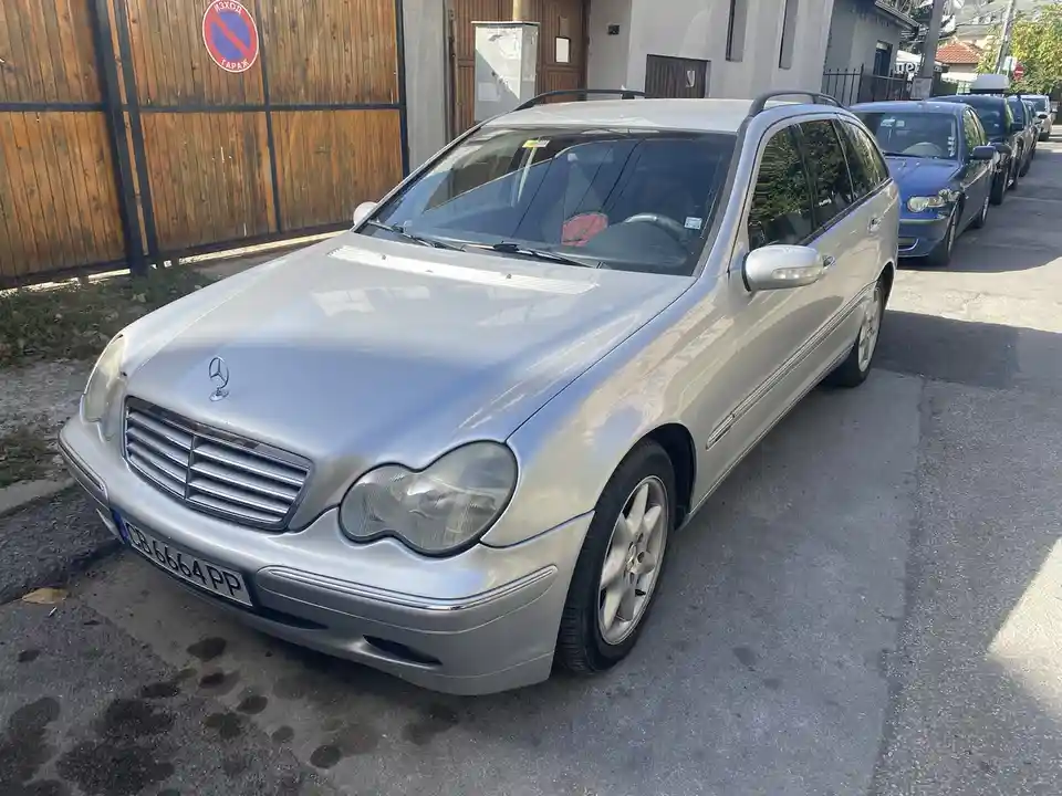 A silver Mercedes-Benz station wagon is parked on a street next to a wooden fence with a red and blue circular sign that…