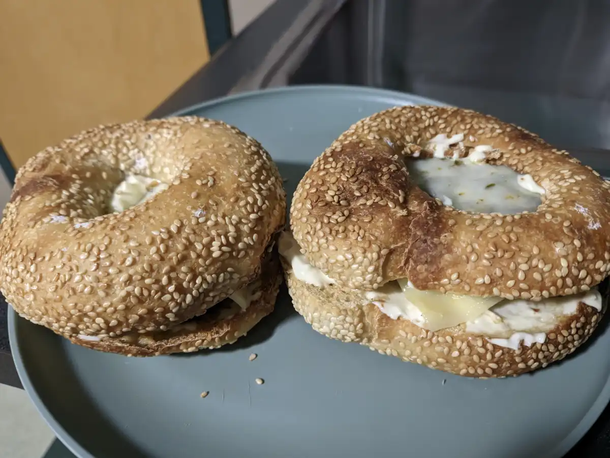 Two sesame seed bagels on a gray plate, one sliced open with cream cheese and a slice of cheese inside, the other whole with…