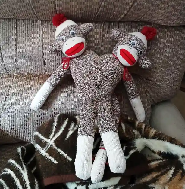 Two identical brown and white sock monkey plush toys are positioned back-to-back on a textured brown couch, with a patterned…
