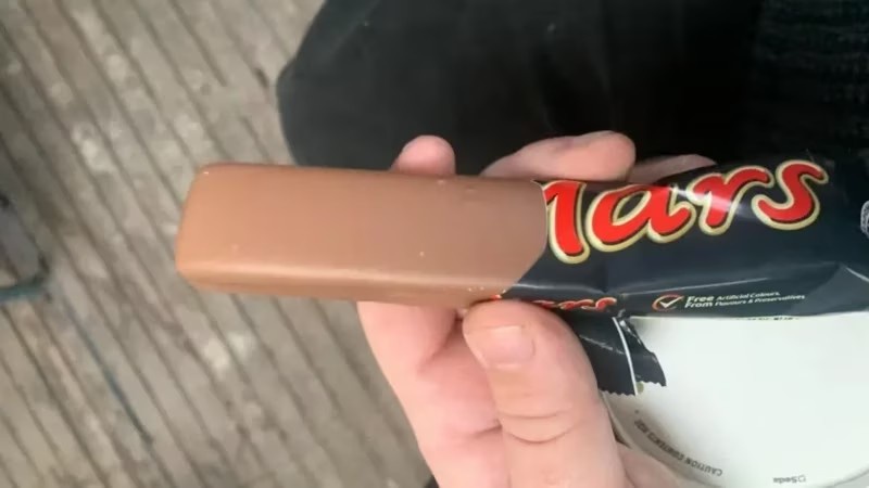 A hand holds a partially unwrapped Mars chocolate bar, with the wrapper visible behind it. The text 'mars' is overlaid in…