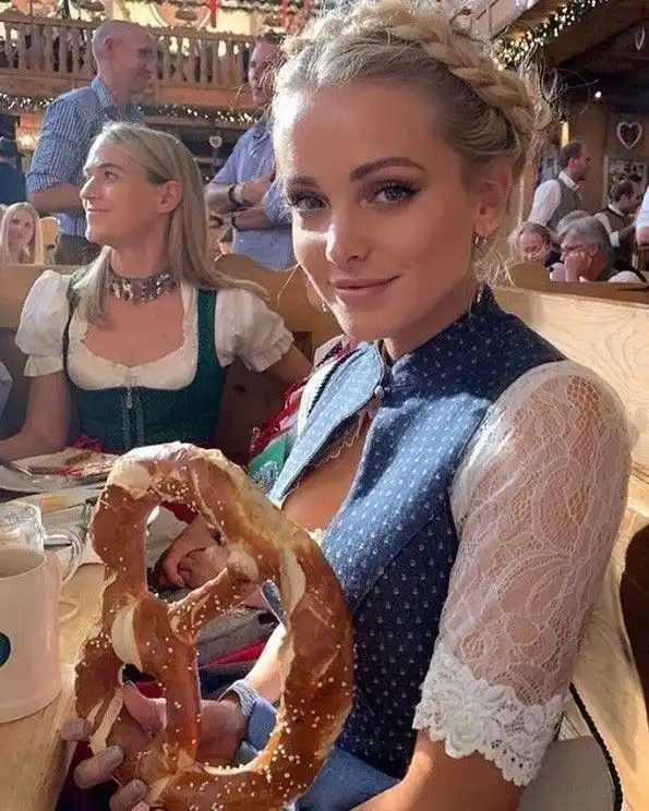 A woman in a traditional German-style dress with lace sleeves and braided hair sits at a wooden table in a crowded…