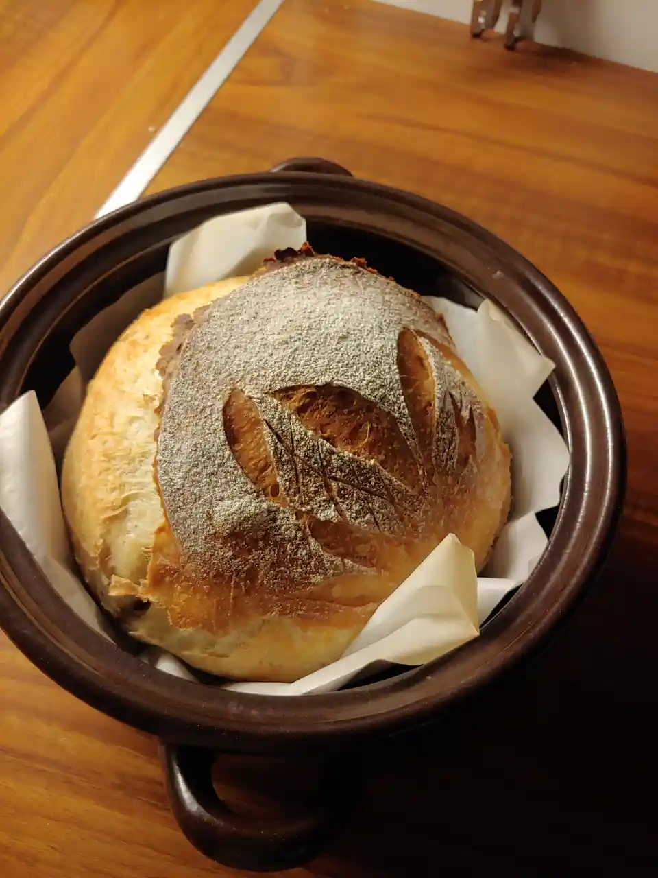 A round, golden-brown artisan bread loaf sits inside a dark brown ceramic bowl lined with white parchment paper, resting on…