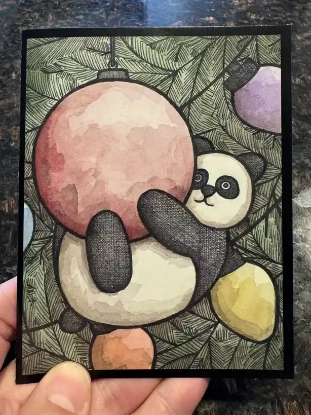 A hand holds a watercolor illustration of a cartoon panda bear peeking out from behind a large pink Christmas ornament…