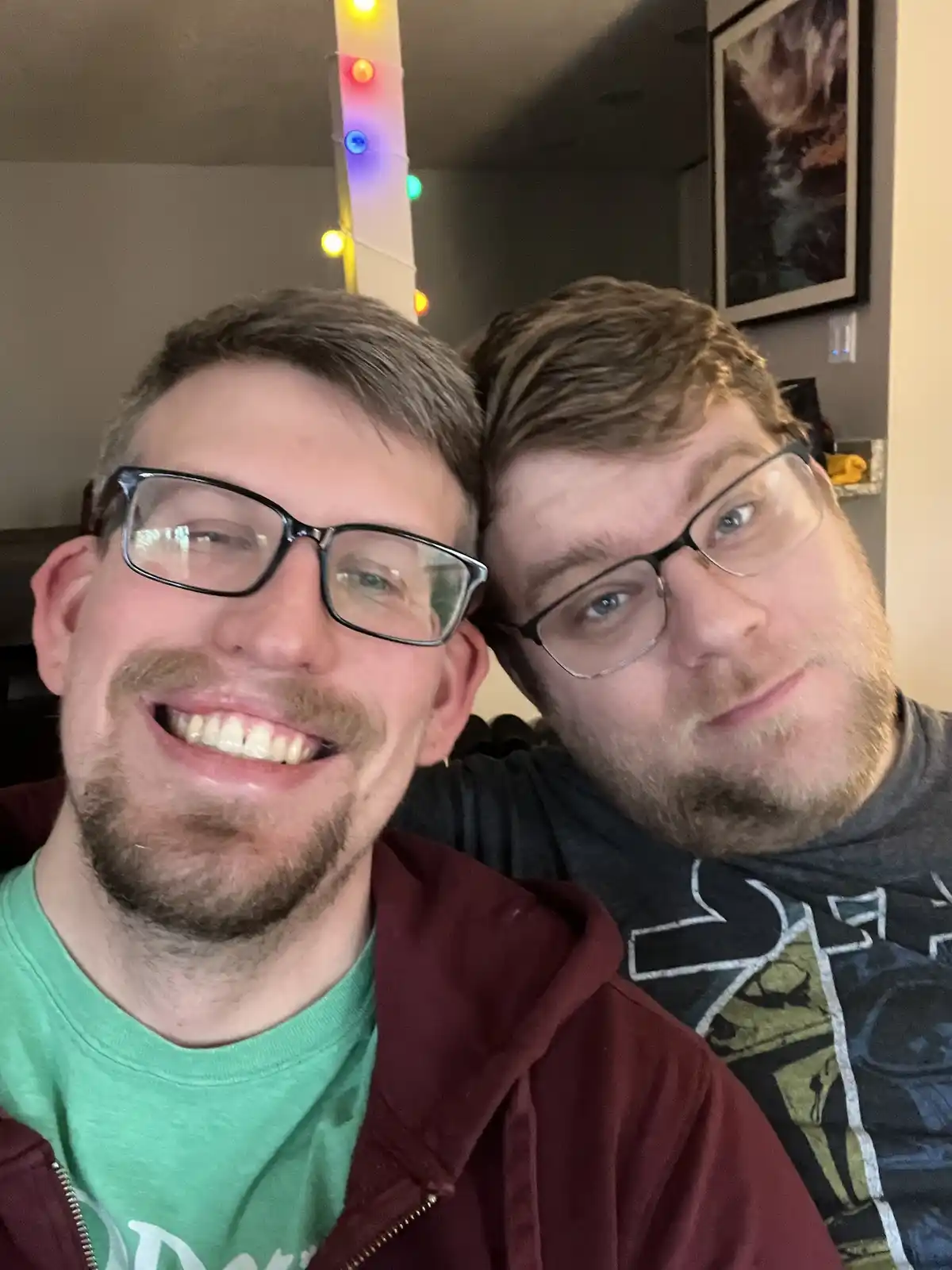 Two men with glasses are in a selfie, one smiling broadly while the other has a subtle smirk. The man on the right has a…