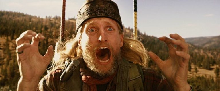 a man with long blonde hair, a patterned hat, and a beard is suspended on ropes in a forested mountainous area, mouth wide…