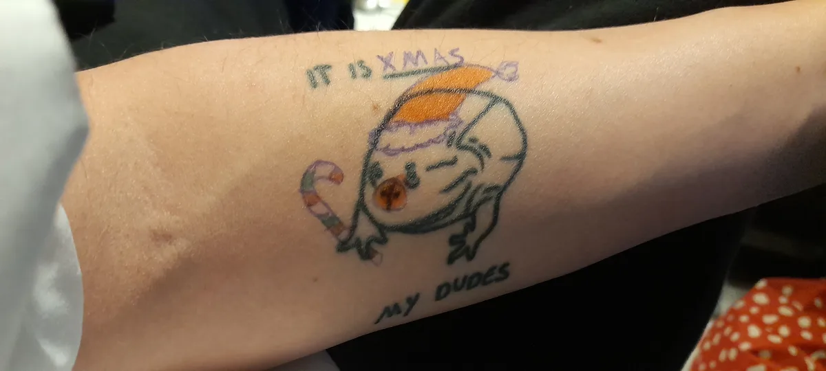 A close-up of a person's forearm showing a freshly inked tattoo of a cartoonish snowman wearing a Santa hat and holding a…