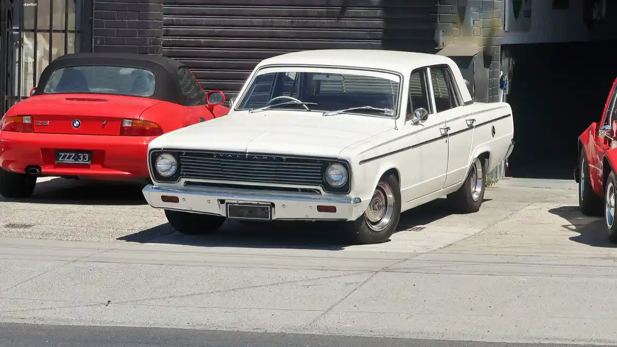 A white 1960s Holden Valiant sedan is parked between a red BMW Z3 convertible and another red car, with the license plate…