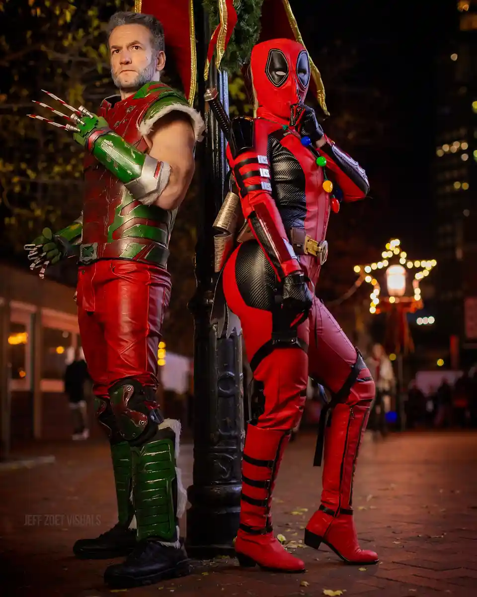 Two people in elaborate costumes stand back-to-back on a city street at night. The person on the left wears a red and green…