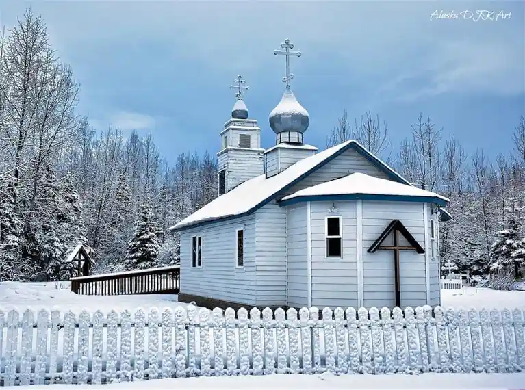 A serene winter scene shows a small, light-blue wooden church with snow-covered onion domes and a cross, surrounded by…