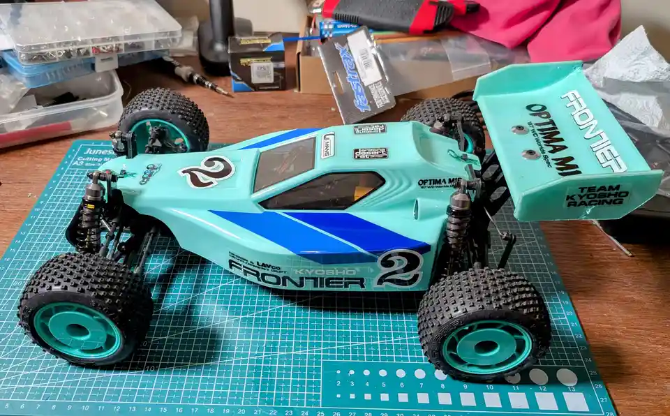 A teal and blue Kyosho Frontier 2 radio-controlled buggy sits on a green cutting mat, surrounded by hobby tools and parts…