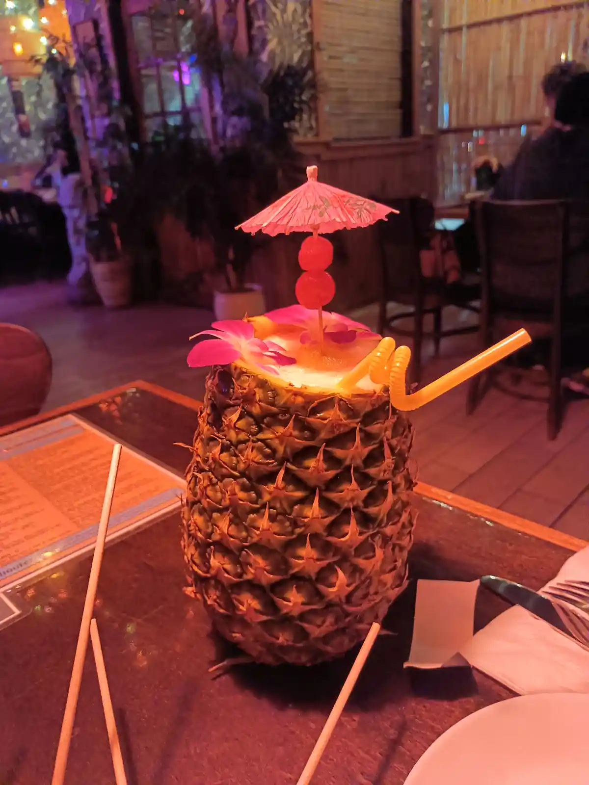 a tropical cocktail served in a hollowed-out pineapple sits on a table in a dimly lit restaurant, adorned with a pink paper…