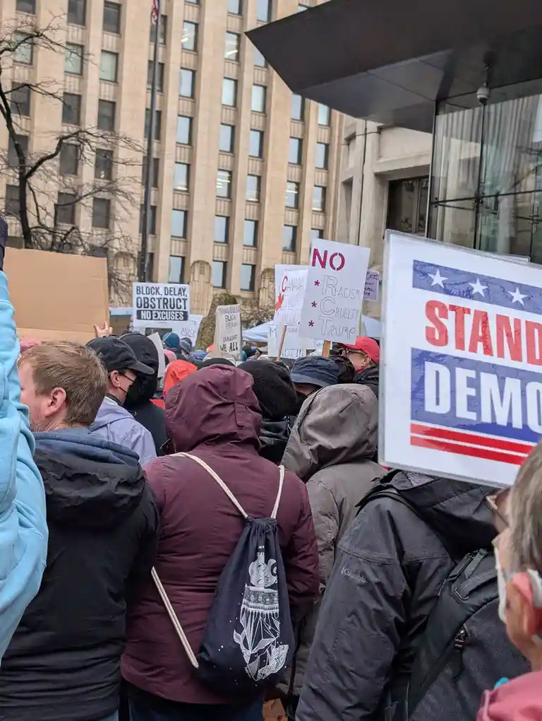 A crowd of protesters at an outdoor demonstration, many wearing hooded jackets and holding signs. Visible signs include 'NO…