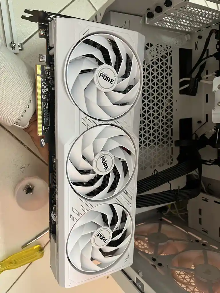 A white Sapphire Pure graphics card with three fans is being held inside a computer case, with a hand visible holding it…