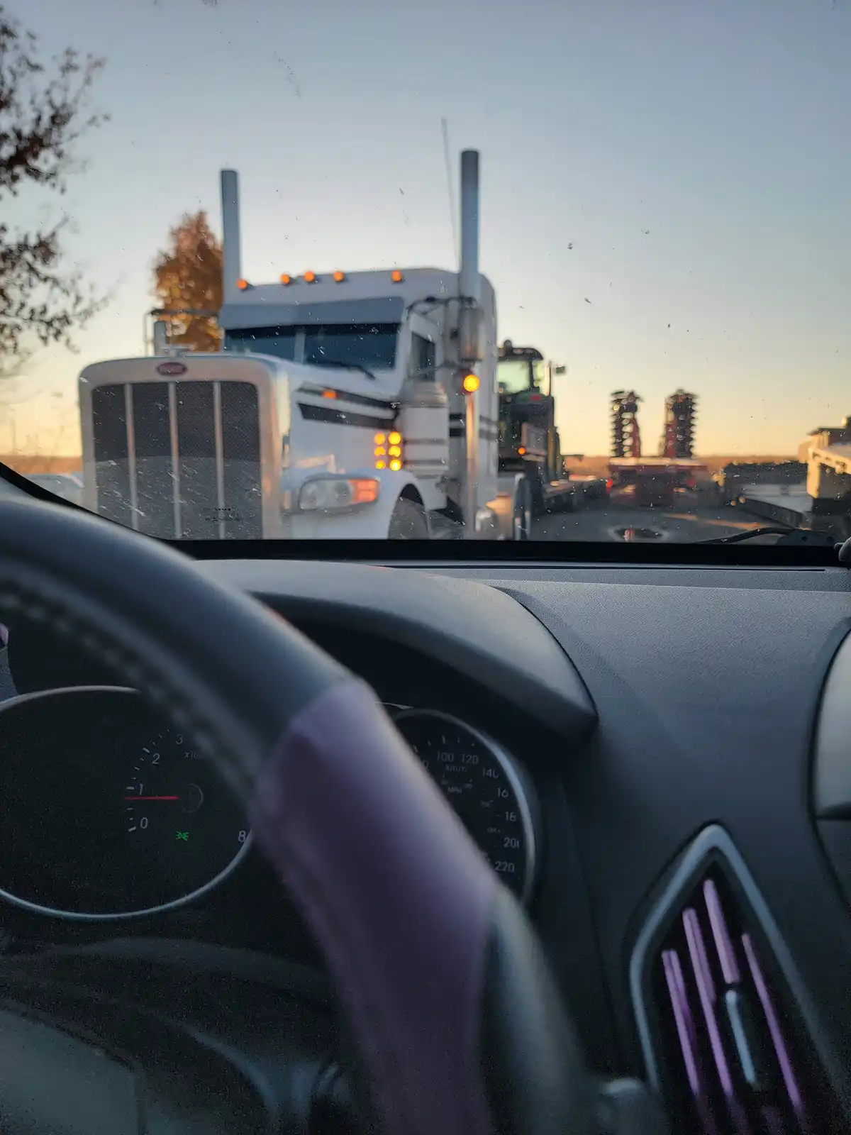 A photo taken from inside a car shows a large white semi-truck directly ahead, with the car's dashboard and steering wheel…