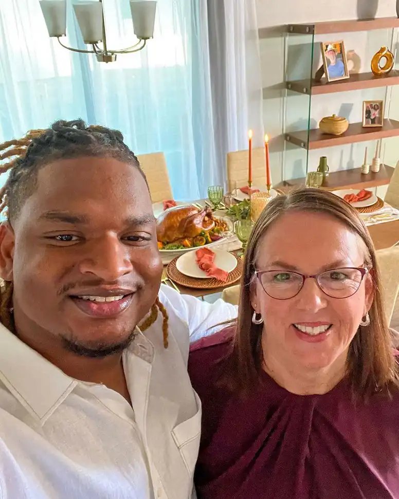 A selfie of a smiling Black man with braided hair and a smiling white woman with glasses, both looking at the camera. Behind…