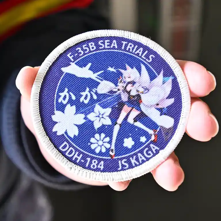 A hand holds a circular blue military patch with white trim. The patch features an anime-style girl with white hair and…