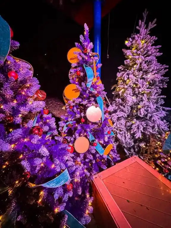 A nighttime photo shows several artificial Christmas trees with purple, frosted needles, decorated with warm string lights…
