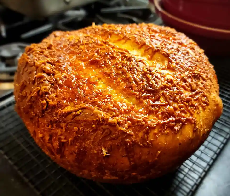 A round, golden-brown baked good, likely a cake or bread, sits on a black wire cooling rack. Its surface is covered in a…