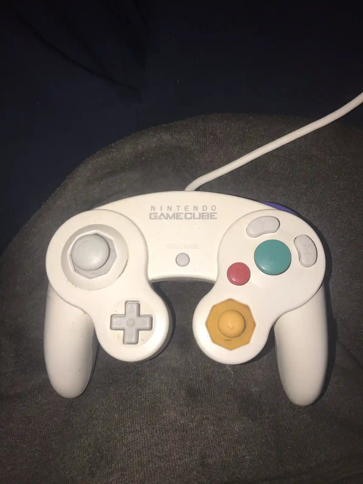 A white Nintendo GameCube controller rests on a dark gray fabric surface, showing signs of wear. The controller has its…