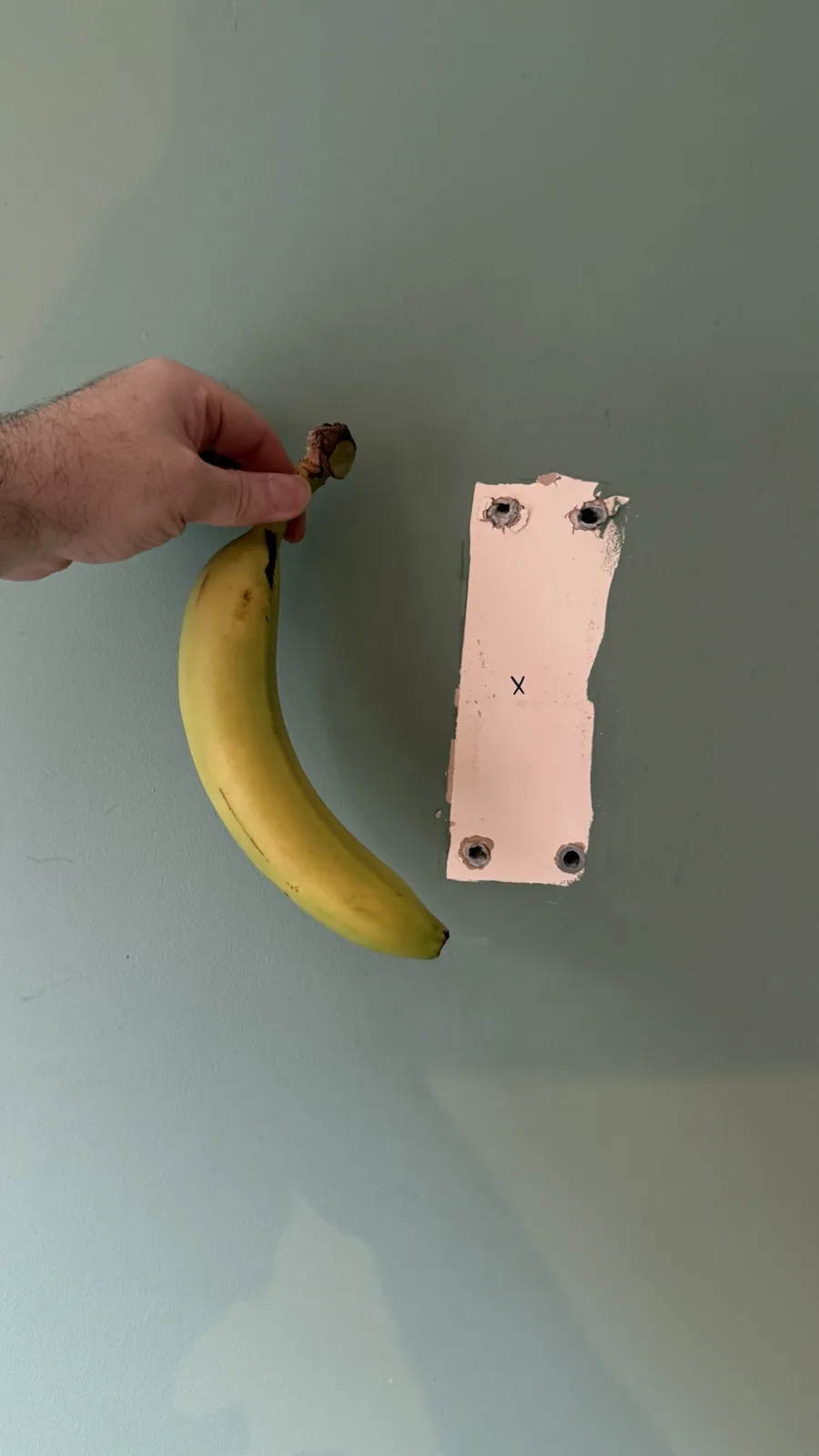A hand holds a ripe yellow banana by its stem, positioning it next to a rectangular patch of wall where a light switch plate…