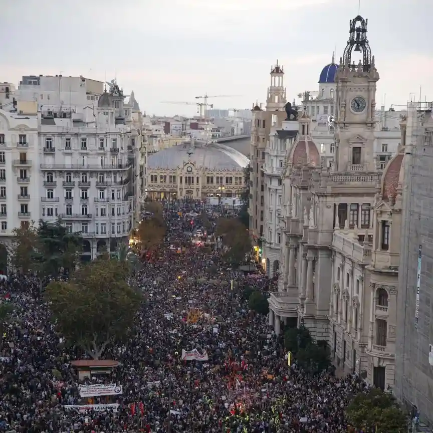An aerial view of a massive protest filling a wide city street between ornate European-style buildings, with crowds holding…