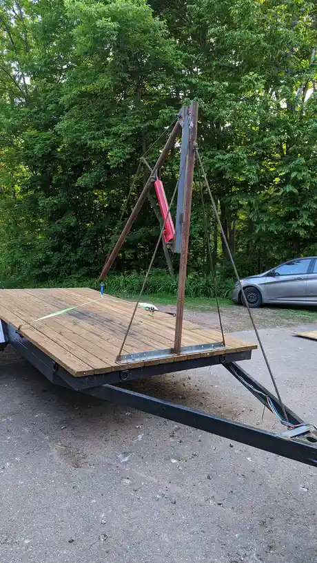 A flatbed trailer with a wooden deck is modified with a crude, homemade crane structure made from rusty metal poles, ropes…