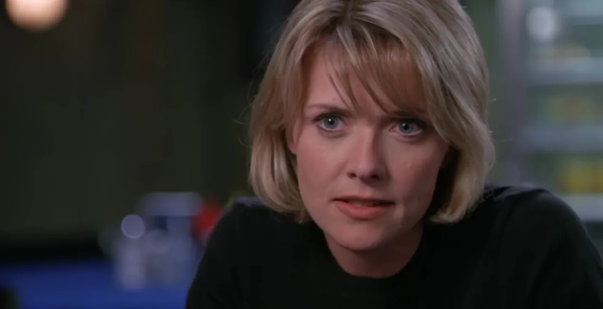 a close-up of a woman with blonde hair and blue eyes wearing a black shirt, looking directly at the camera with a serious…