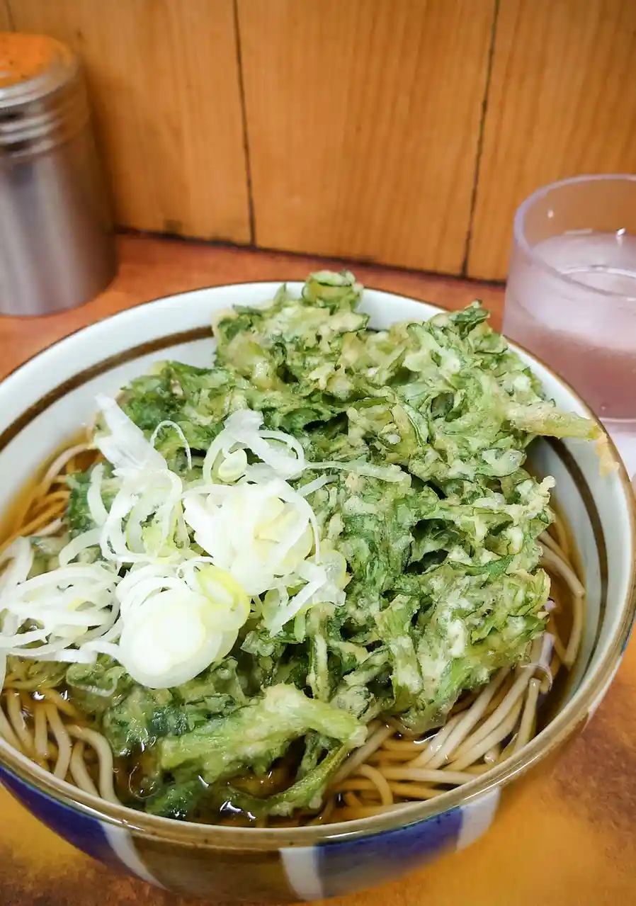 A close-up photo shows a bowl of soba noodles covered with a large, messy pile of tempura-fried green vegetables, likely…