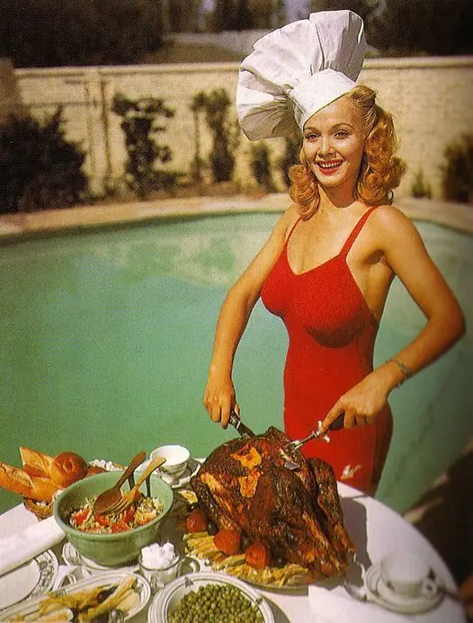 a smiling woman in a red swimsuit and oversized chef's hat stands by a pool, carving a roasted turkey on a table laden with…
