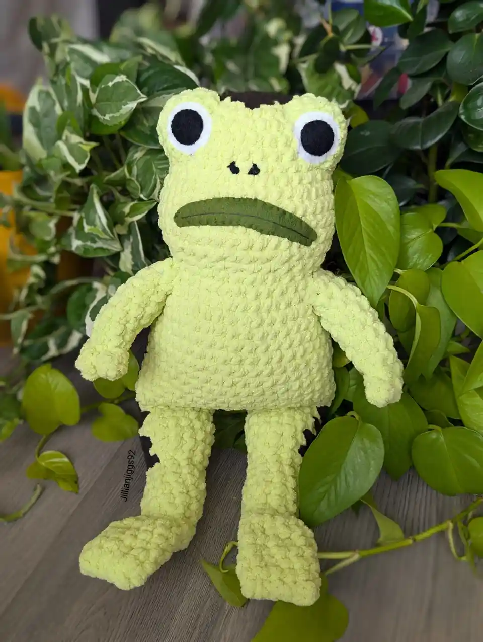 A light green, crocheted frog plush toy with large black eyes and a downturned mouth sits among lush green houseplants on a…