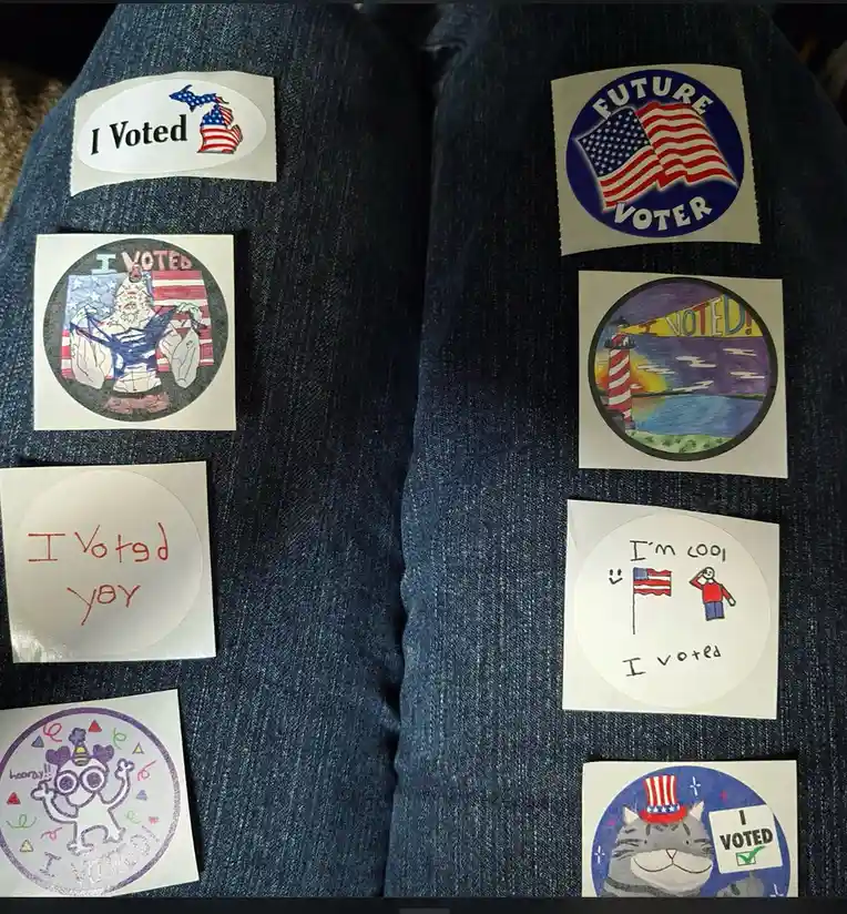 A close-up of a person's denim-clad legs covered in multiple 'I Voted' stickers, including official ones and handmade…