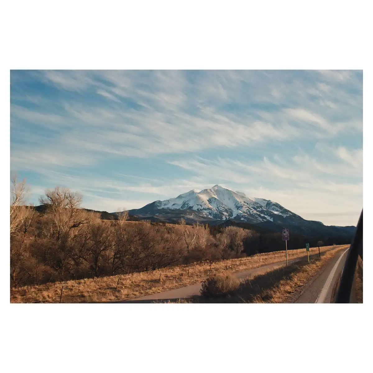 A scenic photo taken from a car window shows a winding road leading toward a snow-capped mountain under a blue sky with…
