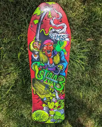 A brightly colored, custom-decked skimboard lies on green grass, featuring a cartoonish pirate with a pink bandana, holding…