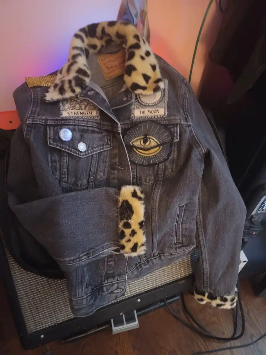 A dark-wash denim jacket with leopard-print faux fur trim on the collar and cuffs, adorned with patches including 'STRENGTH'…