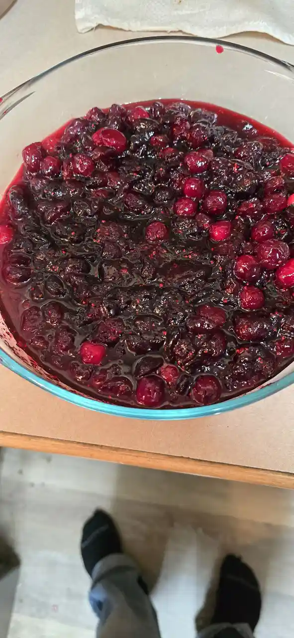 A top-down photo shows a glass bowl filled with dark, glossy cranberry sauce, with whole berries and juice visible. The bowl…