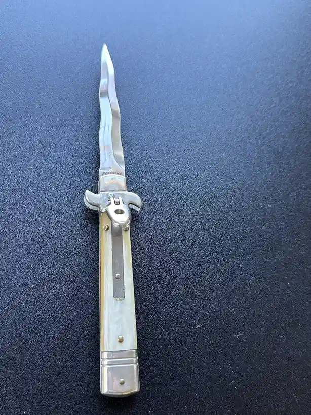 A close-up photo shows a silver-colored folding knife with a wavy blade and a light-colored handle, resting on a dark blue…