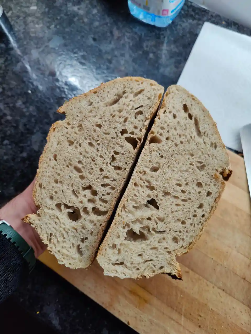 A hand holds two halves of a rustic, artisanal loaf of bread, showcasing its airy, holey crumb structure. The bread rests on…