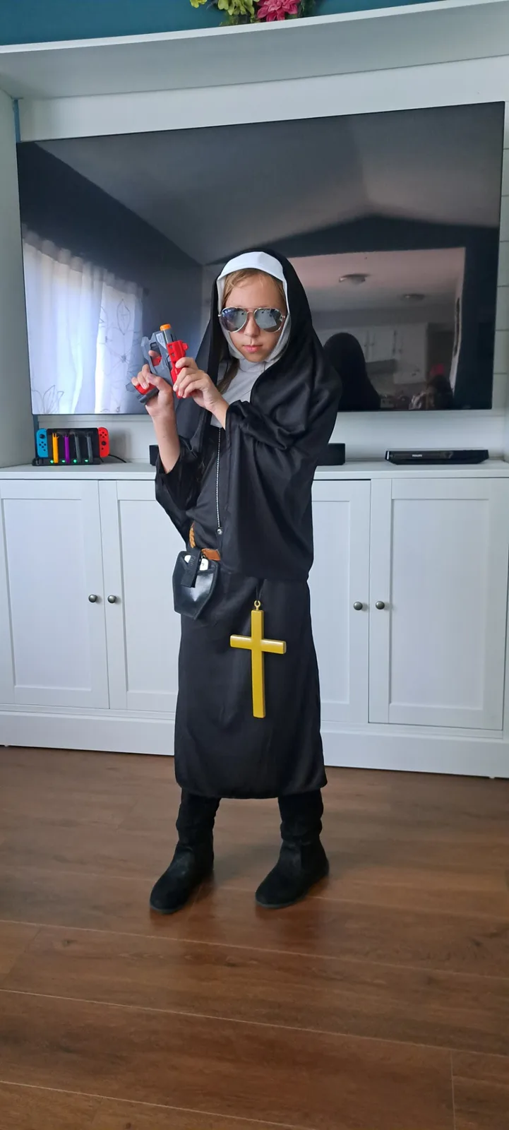 A young person dressed in a black and white nun's habit, wearing aviator sunglasses and holding a red and gray toy gun…