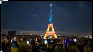 A nighttime photo of a large crowd gathered before the Eiffel Tower, many holding up glowing phones to take pictures. The…