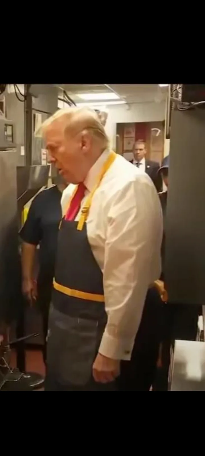 a man with white hair wearing a white shirt, red tie, and a dark blue apron with yellow straps stands in what appears to be…