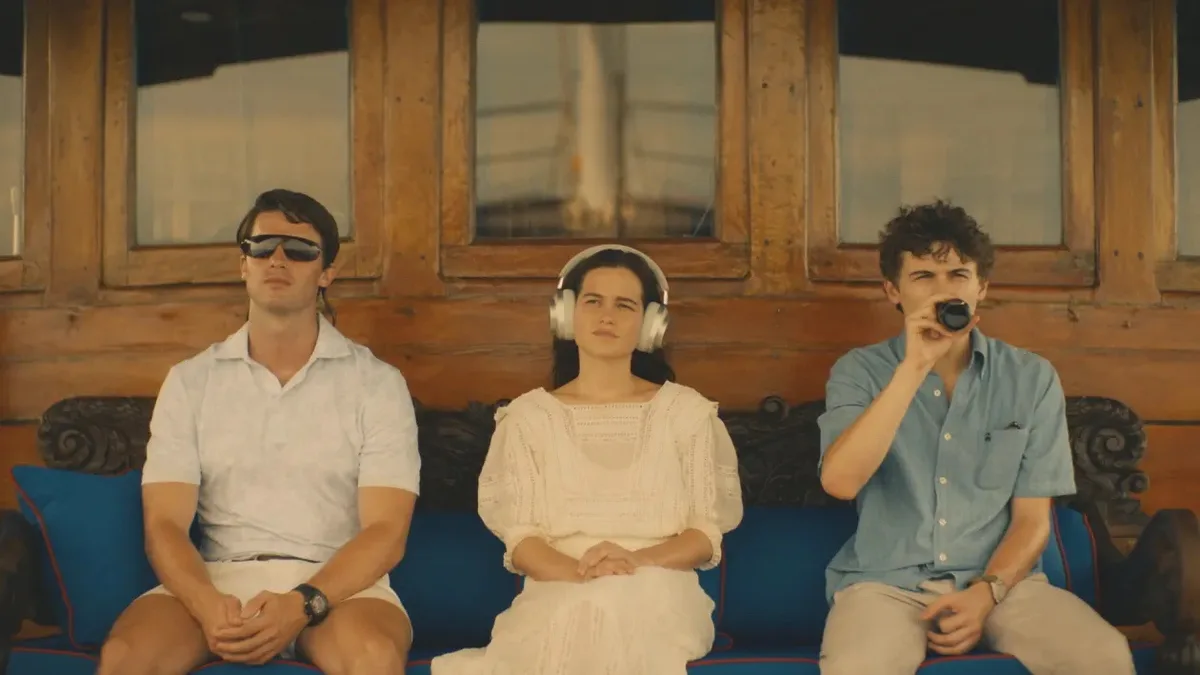 three people sit on a blue couch; the man on the left wears sunglasses, the woman in the middle wears large white…