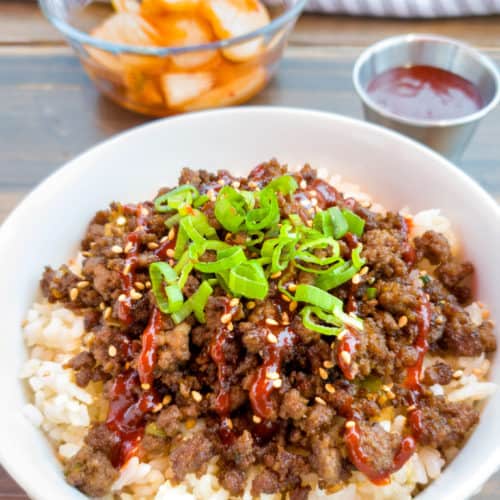 A close-up photo of a white bowl filled with rice topped with browned ground meat, drizzled with red sauce, and garnished…