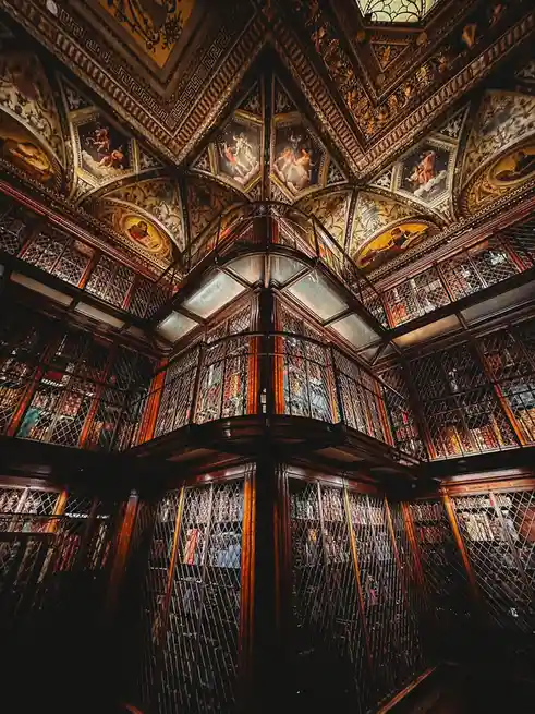 An ornate, multi-level library with dark wood, diamond-patterned glass, and a painted ceiling featuring classical figures…