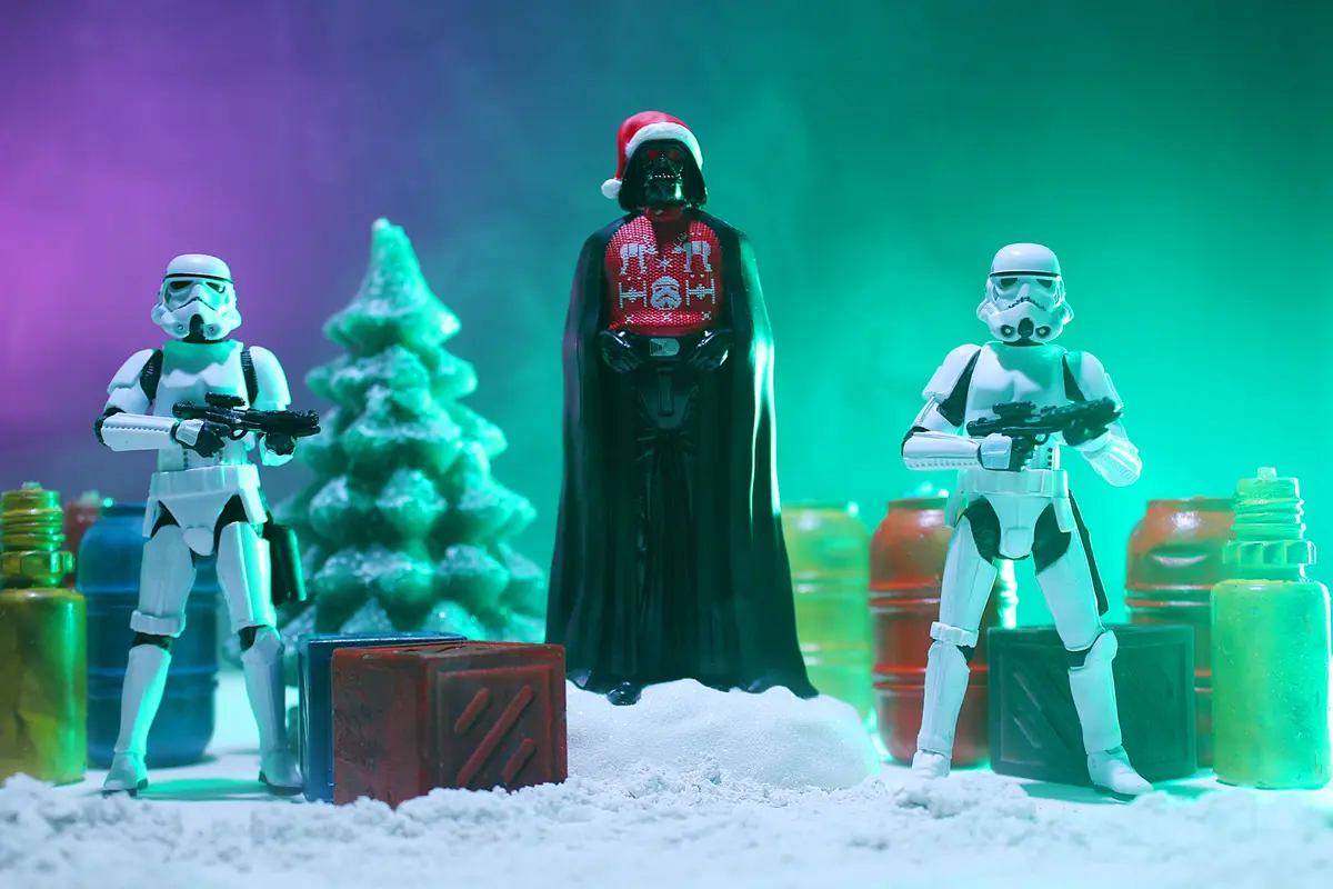 A Star Wars action figure scene features Darth Vader wearing a red Christmas sweater with holiday patterns and a Santa hat…