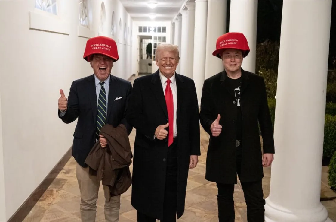 Three men stand in a white-columned hallway, all wearing red 'MAKE AMERICA GREAT AGAIN' hats and giving thumbs-up. The man…