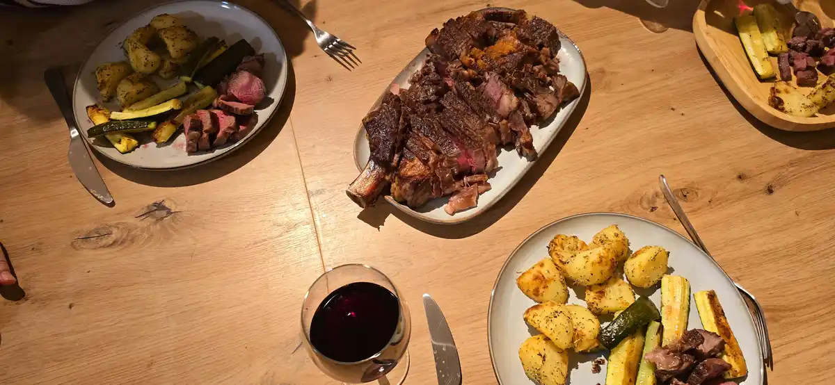 An overhead shot of a wooden dining table with multiple plates of food, including sliced steak, roasted potatoes, and…