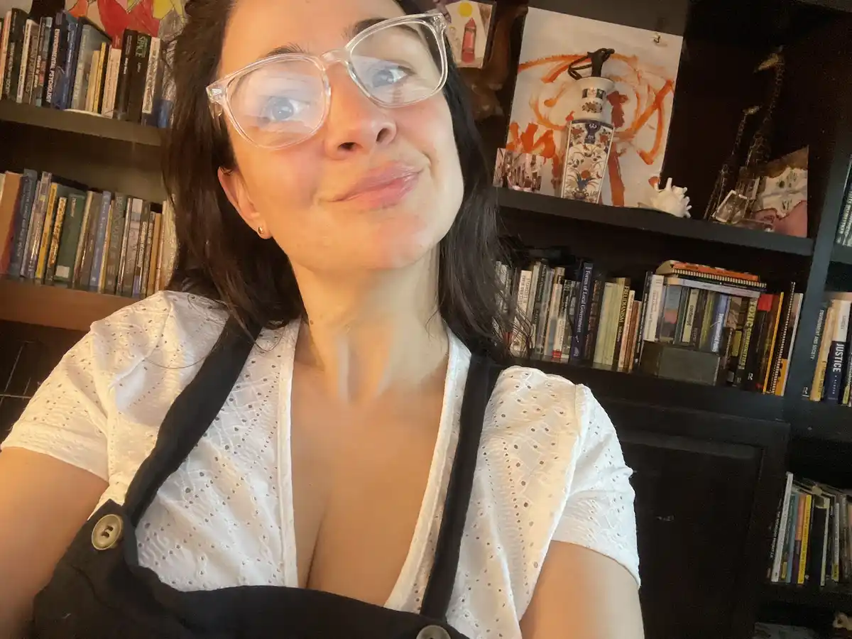 A woman with dark hair and clear-framed glasses looks upward with a smug, self-satisfied smirk while wearing a white eyelet…