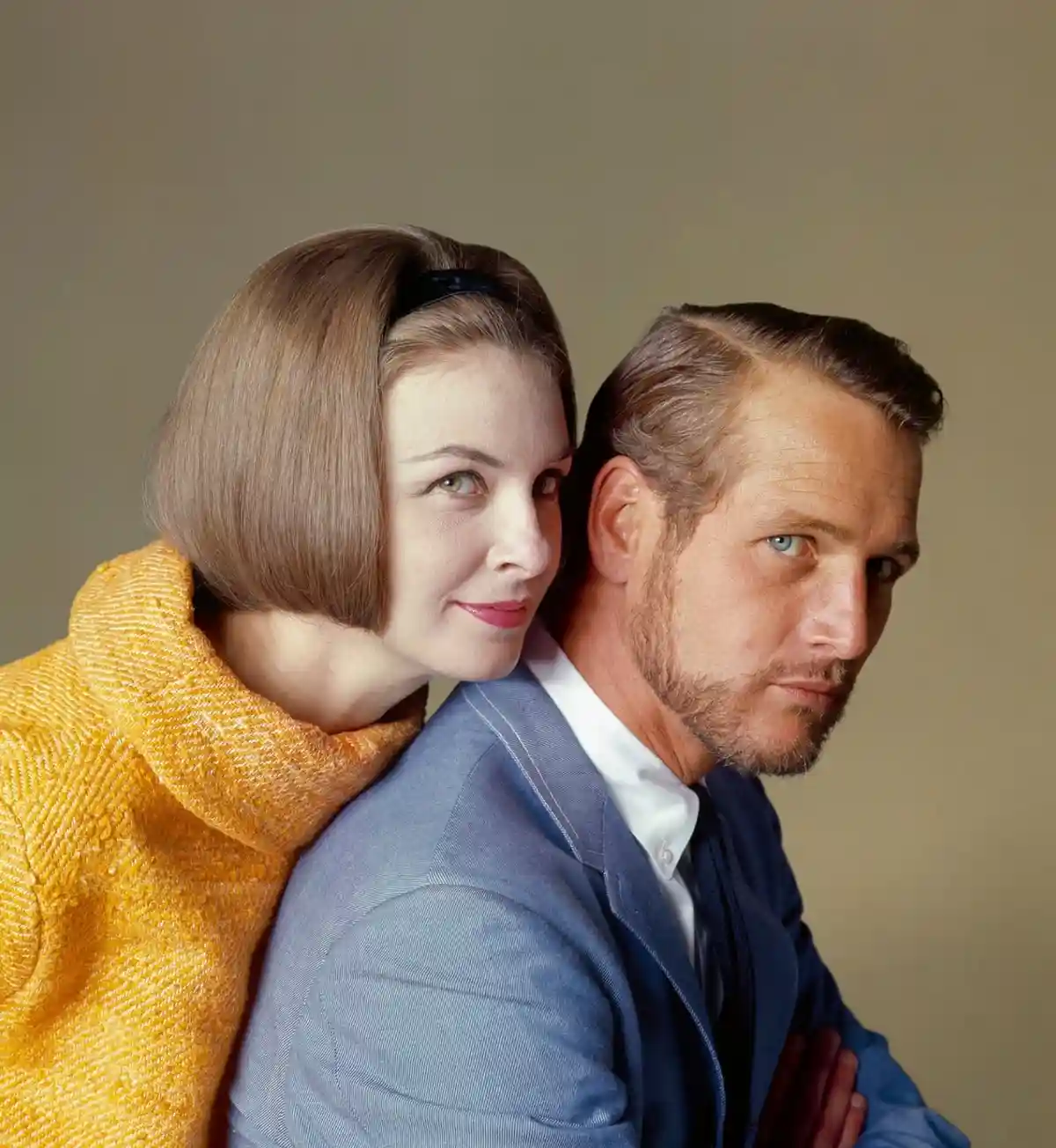 A man and woman are standing back-to-back against a plain background. The woman, wearing a yellow textured coat and a black…