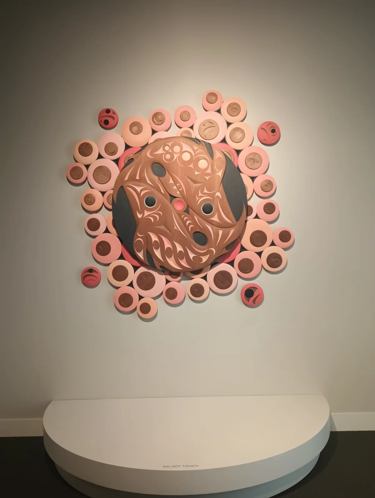 A wooden Indigenous art piece with intricate carvings and a central face surrounded by many smaller circular elements…