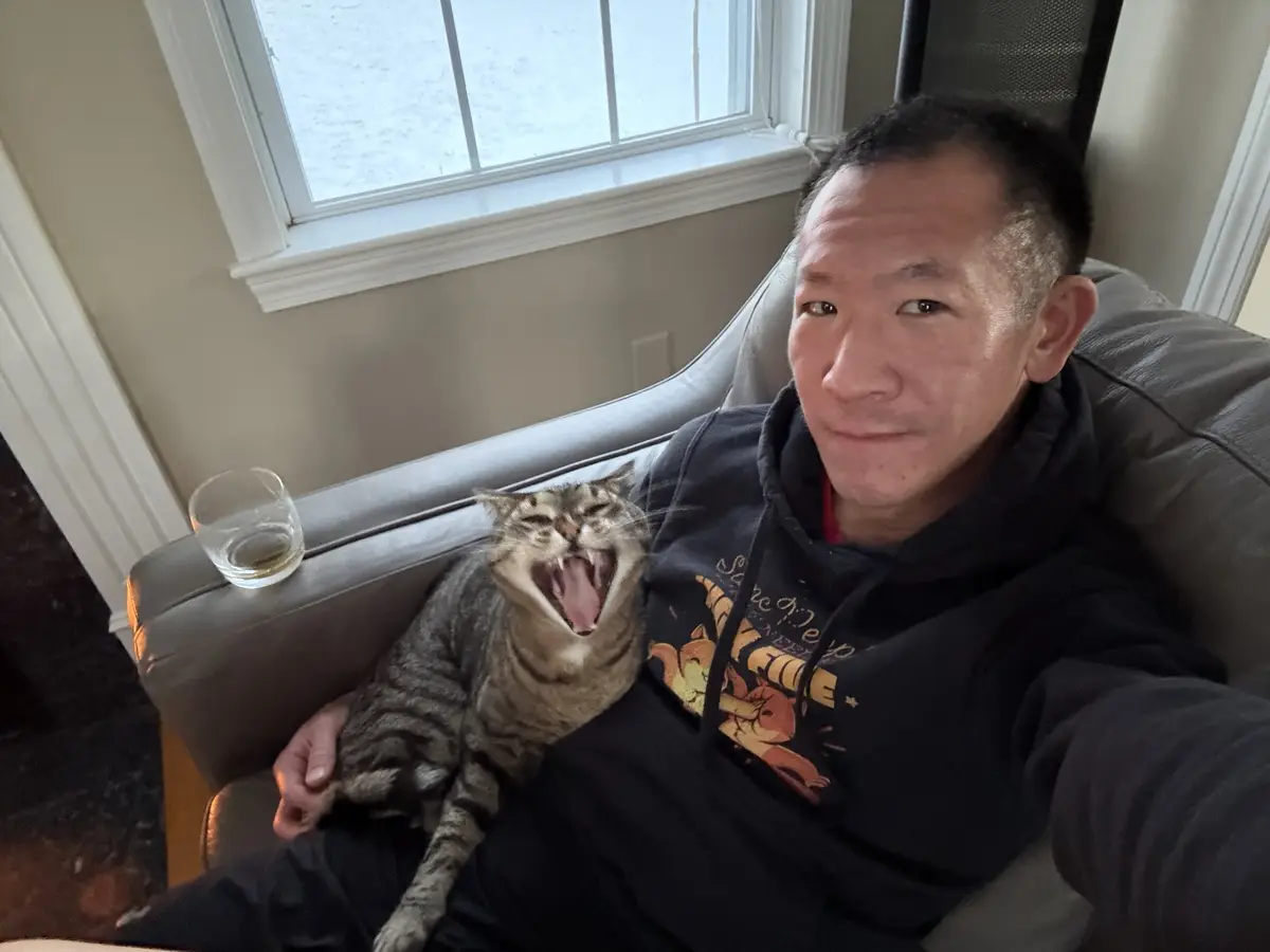 A man in a black hoodie sits on a gray couch, holding a tabby cat that is yawning widely with its mouth open. A glass of…